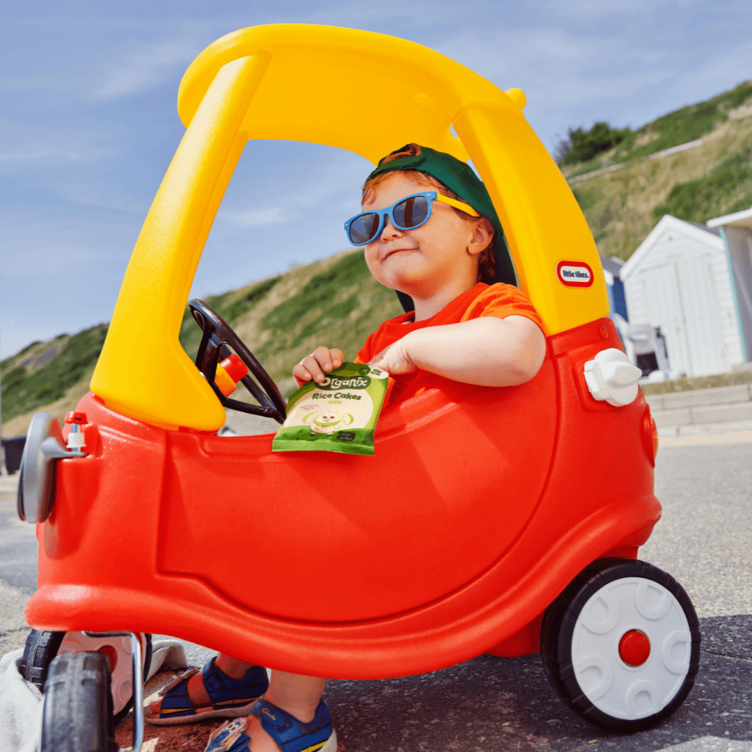 A child holding Organix Rice Cakes in a bright toy car